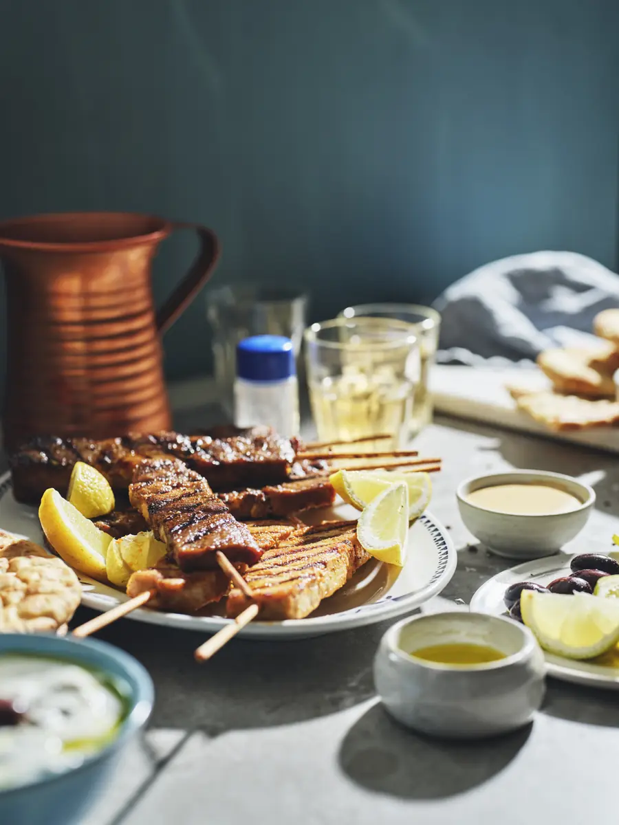 Ett fat med grillade souvlakispett, citron och tzatziki på ett dukat bord