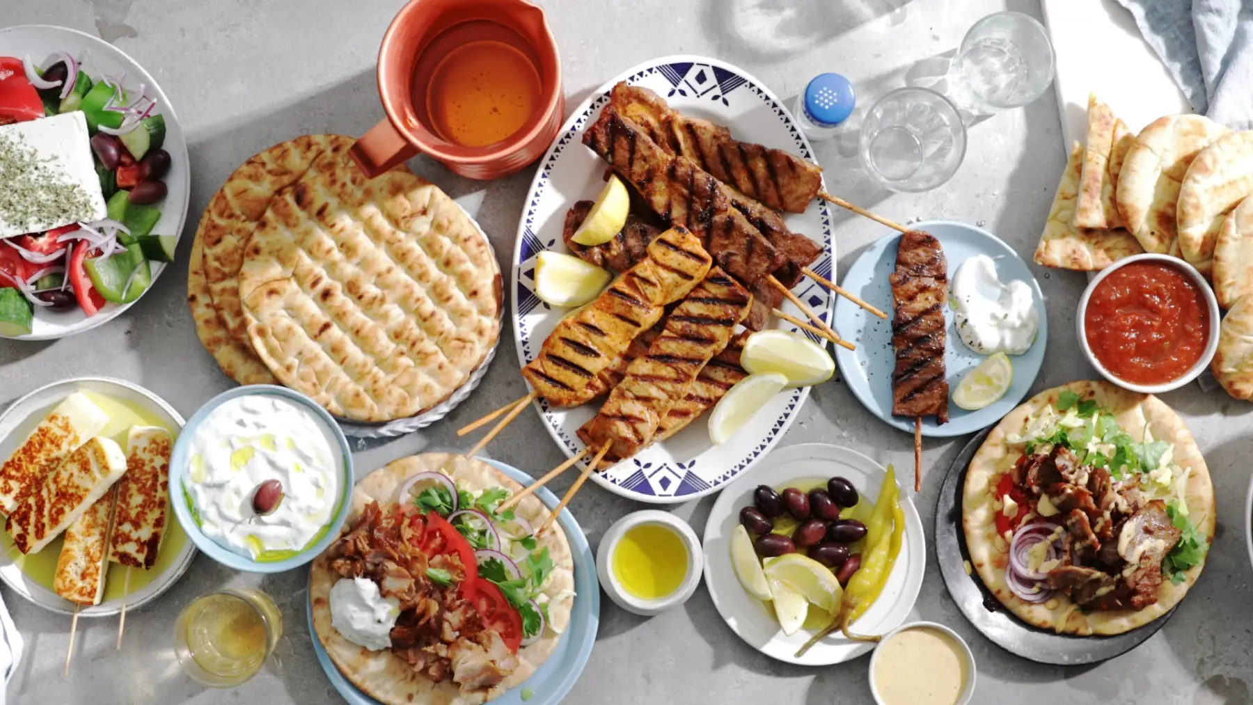Ett dukat grekiskt bord med souvlakispett, pitabröd, tzatziki, oliver och grekisk sallad