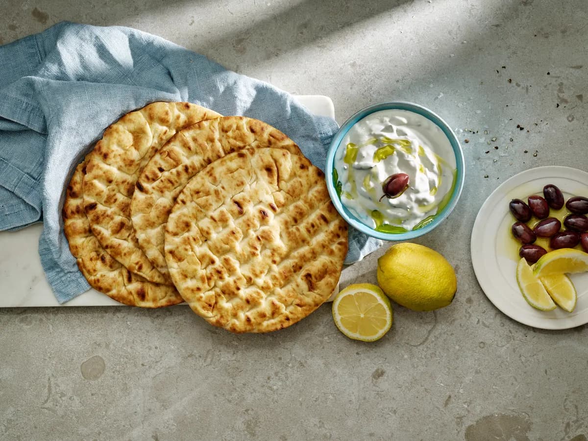 Fyra grekiska pitabröd på en marmorskiva med en skål tzatziki, citron och oliver