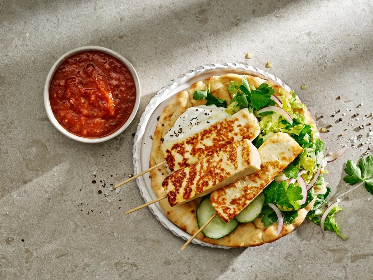Grillostspett på ett pitabröd med strimlad sallad och gurka, bredvid en skål tomatsalsa
