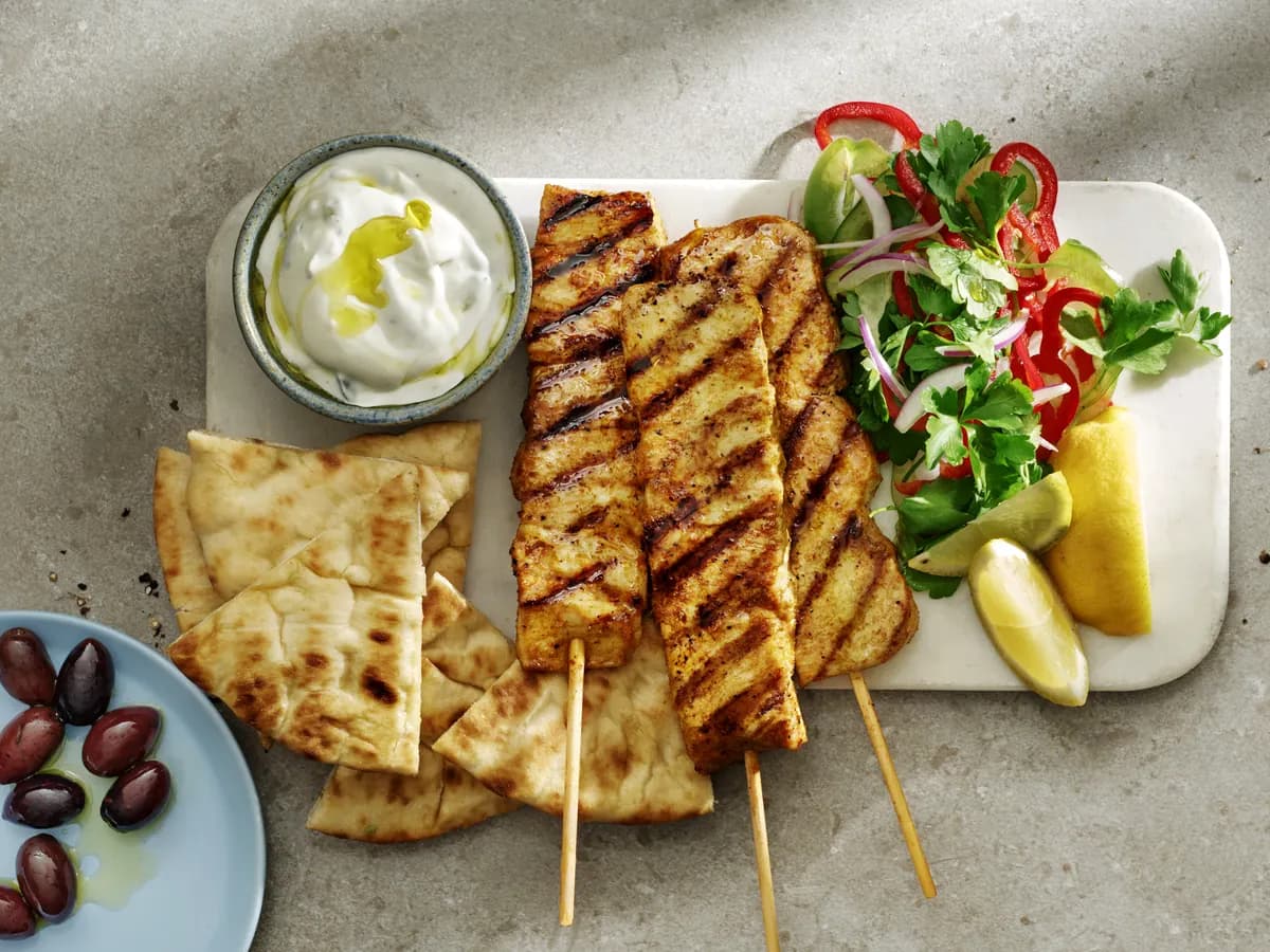 Tre grillade kycklingspett på en marmorbricka med tzatziki, pitabröd i klyftor, sallad med persilja och paprika, citronklyftor och en skål oliver