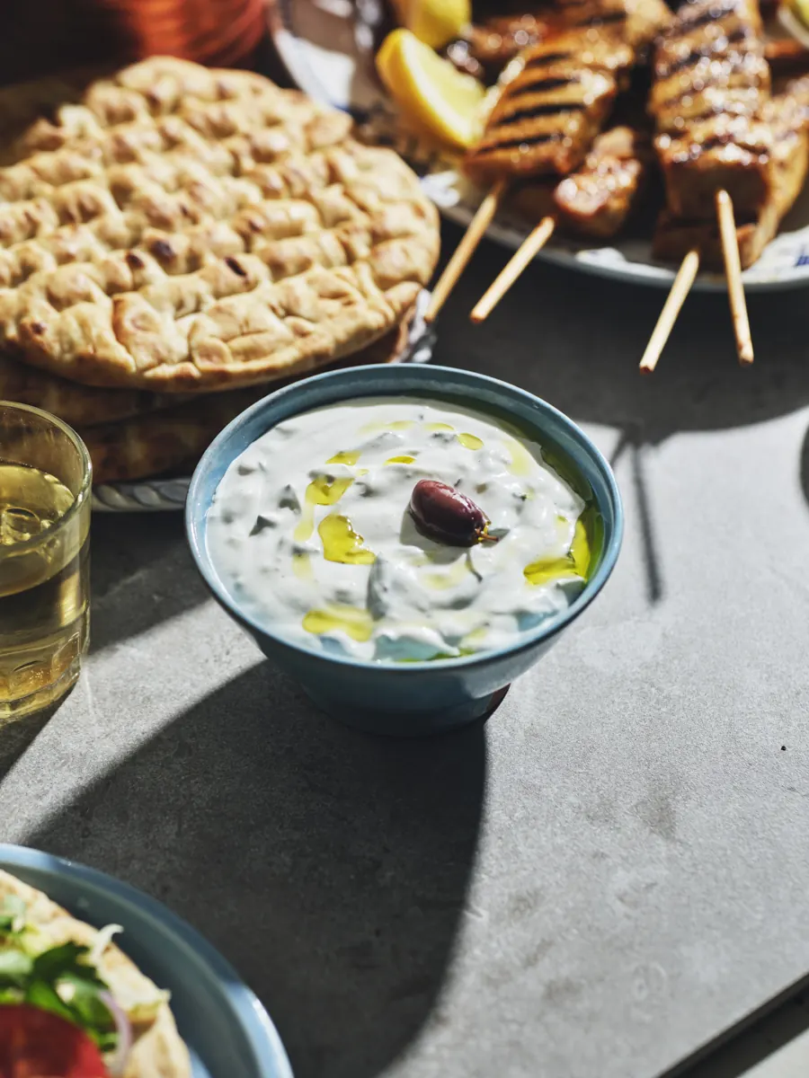Skål med tzatziki toppad med olivolja och en oliv, bredvid en stapel pitabröd